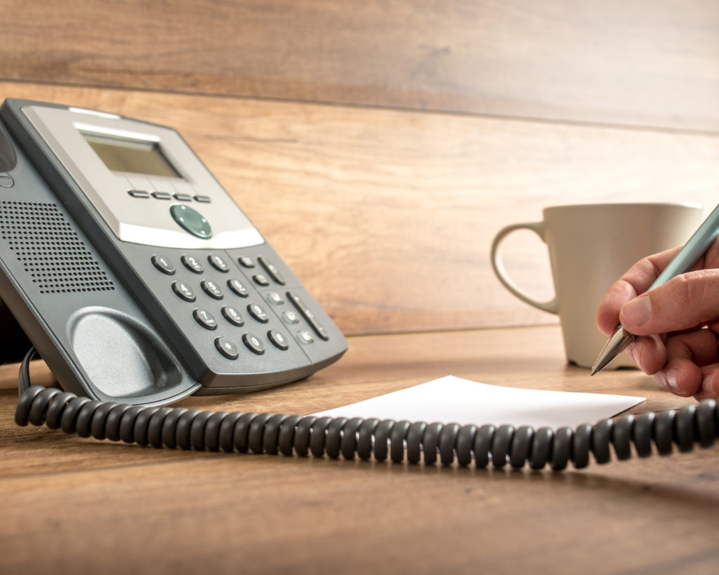 Picture of telephone, phone line, coffee cup, and hand taking notes with pencil