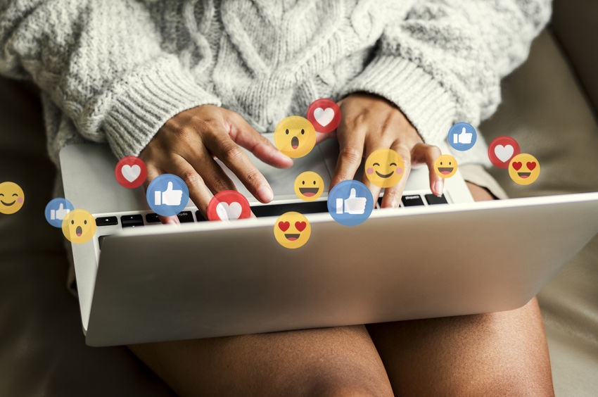 Picture of a woman typing on laptop with social media and emojis around her hands