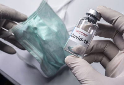 Picture of gloved hand holding bottle marked vaccine Covid-19 and other hand holding ble face mask employScientist holds a coronavirus vaccine, conceptual image