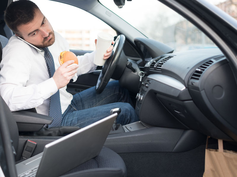 Picture of man in drivers seat of car eating and working on laptop computer