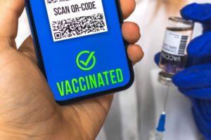 Picture of hand holding phone with Vaccinated checkmarked Scan QR-code next to a blue gloved hand holding a vaccine bottle and needle